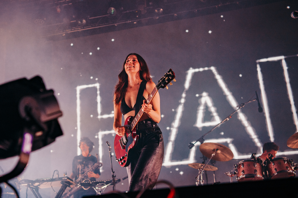 A singer with long dark hair, dressed in a black bikini top and pants holds a red guitar. She is looking out from the stage, two musicians behind her. This is Haim performing at Laneway.