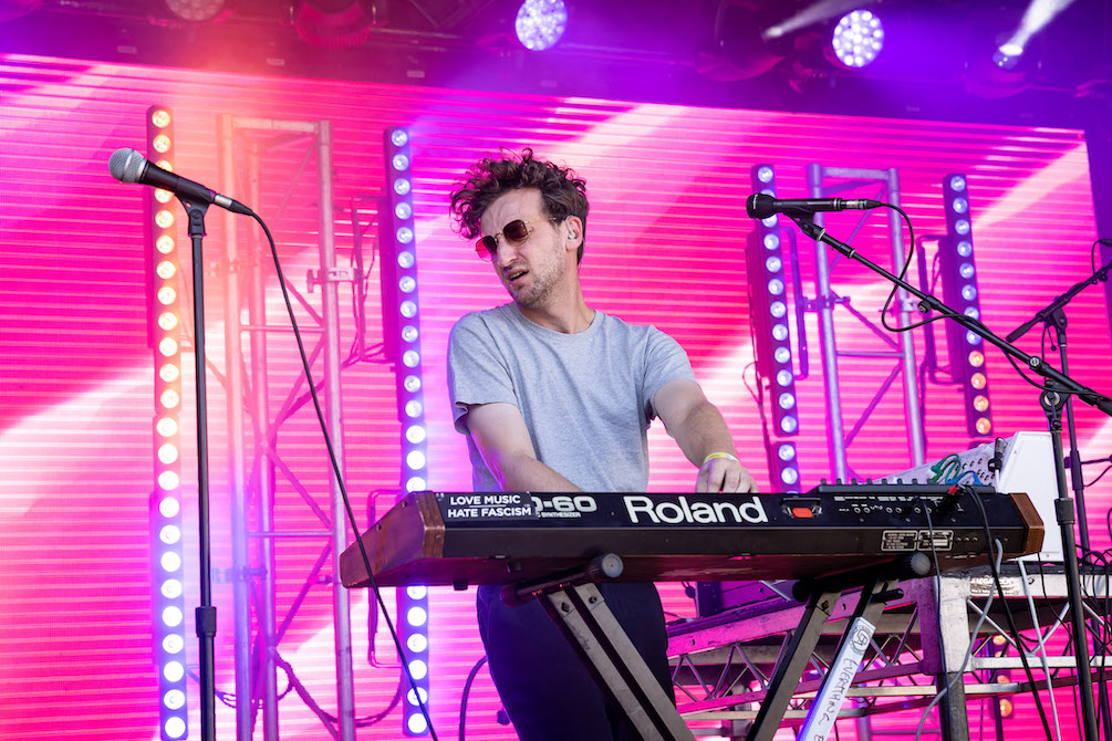 A musician in a grey t-shirt stands behind a Roland keyboard, microphones either side of him. Behind him, the wall is striped bright pink with towers of gold and yellow strobe lights.  It is Harvey Sutherland performing at Laneway.