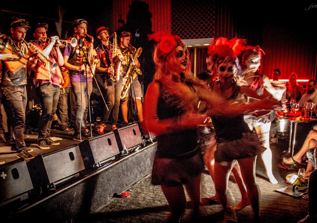 Two people in short skirts wearing headgear and masked make-up dance in front of a colourfully dressed brass band. They are enjoying Fremantle Carnevale.
