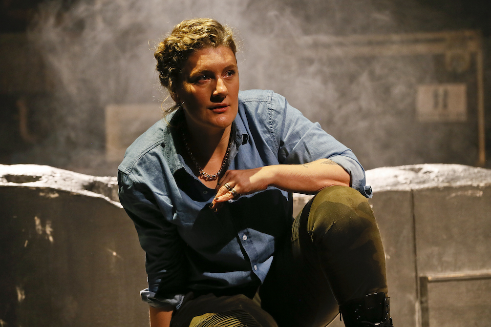 A blonde woman in a denim shirt and camouflage pants squats, one elbow resting on her knee. She is wearing a beaded necklace and rings. Behind her is a brick wall and a smoky haze. This is Virginia Gay in Cyrano.