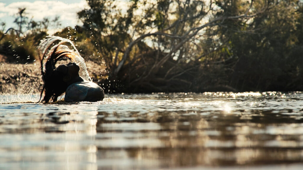 a promotional image from 'Punkaliyarra' by Big hART, showing a woman in a water hole, flicking her hair back.