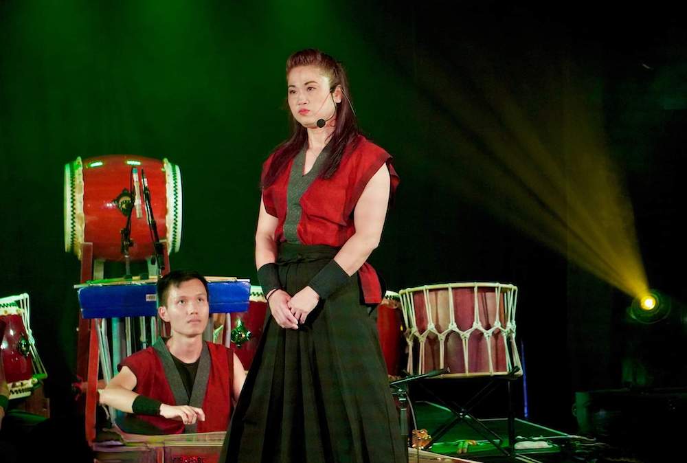 A young woman from Taiko On stands in from of a drummer, with drum kits in the background. She wears a red kaftan style top and a black skirt.