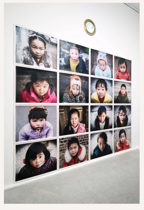 A work from Beijing Realism shows 16 portraits of Chinese children looking straight at the camera. They are dressed warmly for cold weather.