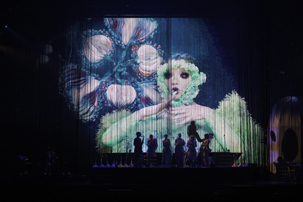 Musicians on stage are dwarfed by a fantasy-like image projected onto a large screen. A fairy-like creature with large eyes and flowers surrounding her face holds her hands to her face as if in surprise. Behind her are dreamy blue and pink flowers. This is Bjork's Cornucopia.