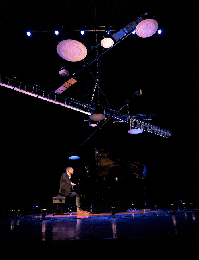 A large sculptural mobile with different shaped lights suspends from the ceiling. Below Cedric Tiberghien sits at the piano, a blue light above his head.