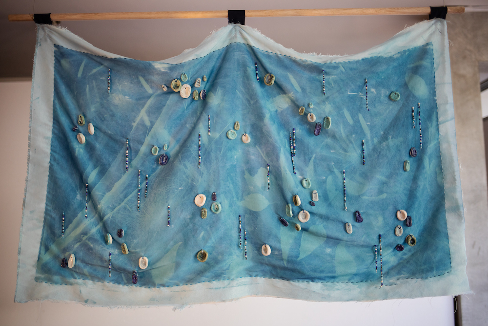 A photo from Know Thy Neighbour #3, of an artwork made of fabric, dyed cobalt blue with aqua leaf shapes and shells and beads sewn in little clusters.