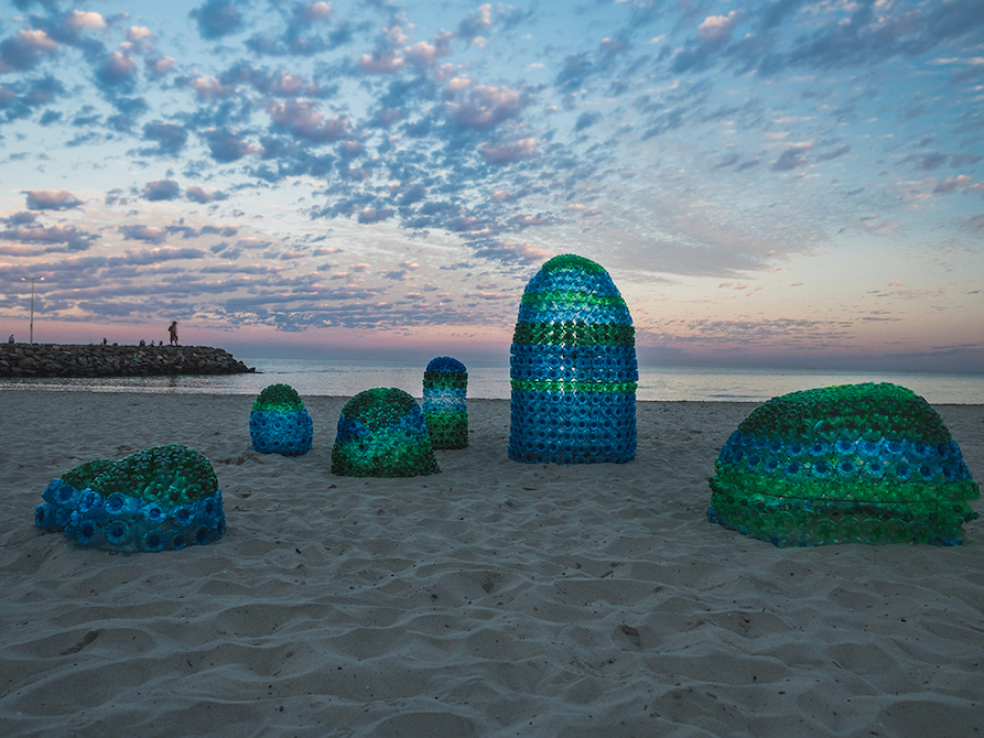 Sea-green sculptures, some shaped like bullets, others like pods, dot the sand. In the background, a dusky pink  sky glows over the ocean and groyne.