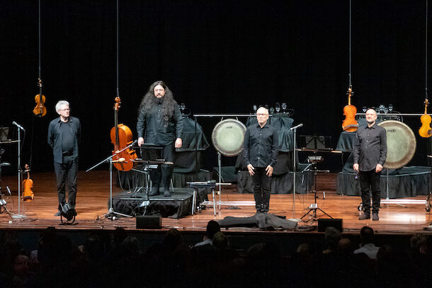 Four men in black stand on a stage, arms by their sides. Their string instruments hang from the ceiling next to them. This is Kronos Quartet preparing to perform 'Black Angels' at Perth Festival.