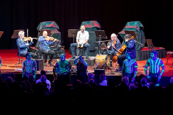 Noongar artist Maatakitj sits on a stage surrounded by four musicians playing stringed instruments. In front of them are Noongar dancers painted in traditional stripes and markings. This is Kronos Quartet's concert at Perth Festival, performing the world premiere of Bindari.
