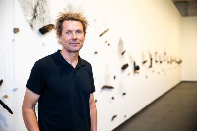 Artist Robert Andrew standing in front of his work Tracing Inscriptions – Moondang Dandjoo Koorliny, which features natural objects on the wall which scrape as they move creating patterns.