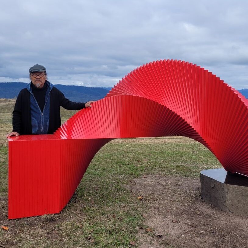 Artist Takeshi Tanabe wears a cap and warm dark clothes. He is standing behind a vibrant red sculpture that is a blend of ridges and curves. He is outside, grey skies behind him.