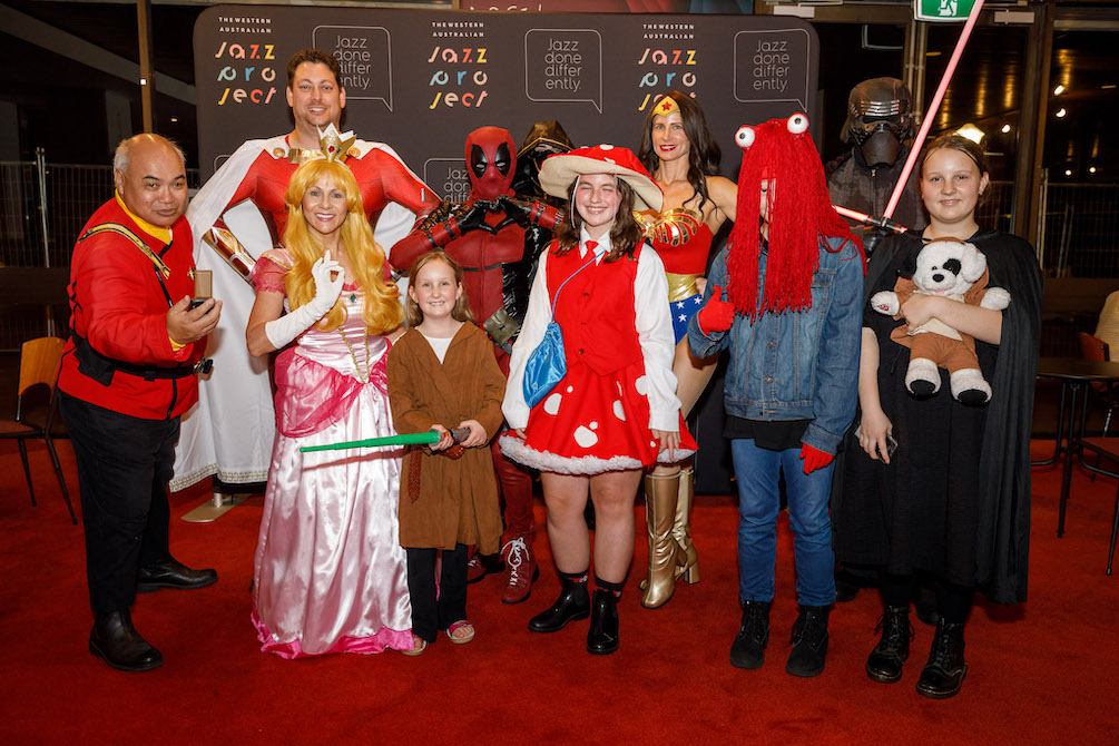 A group of people in cosplay pose for the camera .They range in ages and wear everything from a Wonder Woman outfit to a Jedi suit. They are part of the audience at Western Australia Jazz Project's Heroes, Sci-fi and Sorcery: A Tribute to Comic-con.
