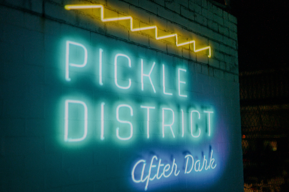 A neon sign that says "PICKLE DISTRICT after dark" glows in the dark.