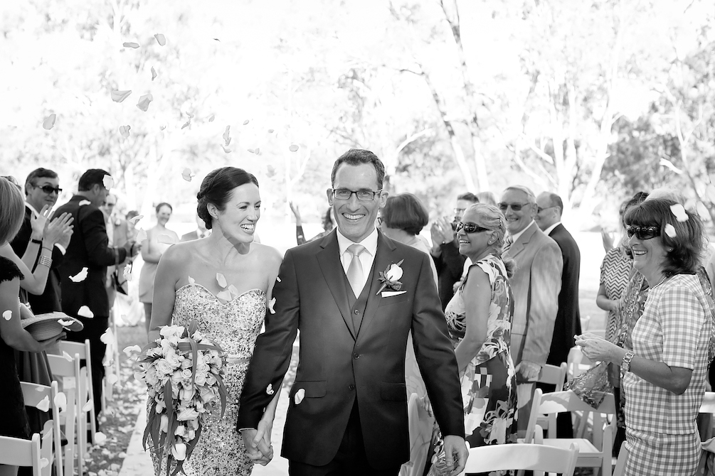 A black and white image of a newly-wed couple striding down the aisle, smiling broadly. She is wearing a strapless gown and carrying a bouquet of flowers, he is in a smart suit and tie, with a corsage on his lapel. Behind them people are standing, smiling and clapping in celebration. This is the wedding of Helen Kruger and Antony Brian.