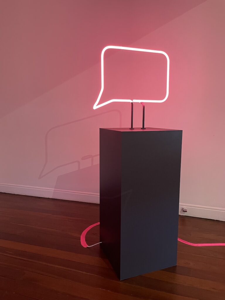 A work from Tracing Absence - a  speech bubble made of pink neon light stands atop a black plinth.