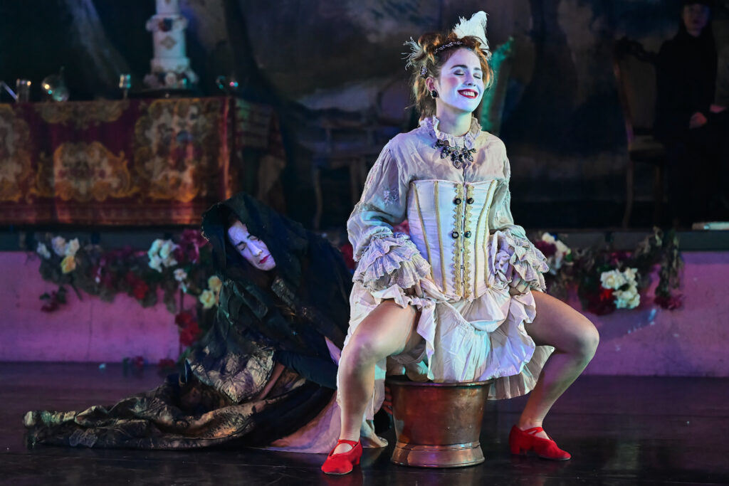 Smiling beatifically, a young woman dressed in an eccentric baroque costume hold up her skirt and squats, while a black clad person hold a chamber pot underneath her.