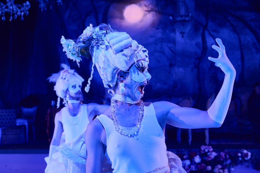 In the foreground a person wearing an extravagant baroque style headpiece and eye mask holds one hand in from of their face, their mouth open as if in a scream. Another person in the background appears to be doing the same gesture.