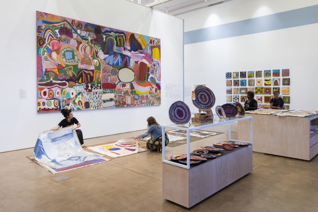 A photograph of the interior of an art gallery filled with colourful paintings and objects. Some people are laying works on the ground.