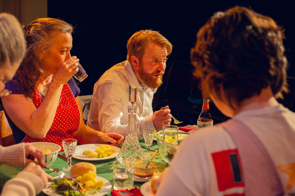 A family sits around a heavily laden dinner table, mid-meal. The focus is on a man with a beard who appears to be speaking.