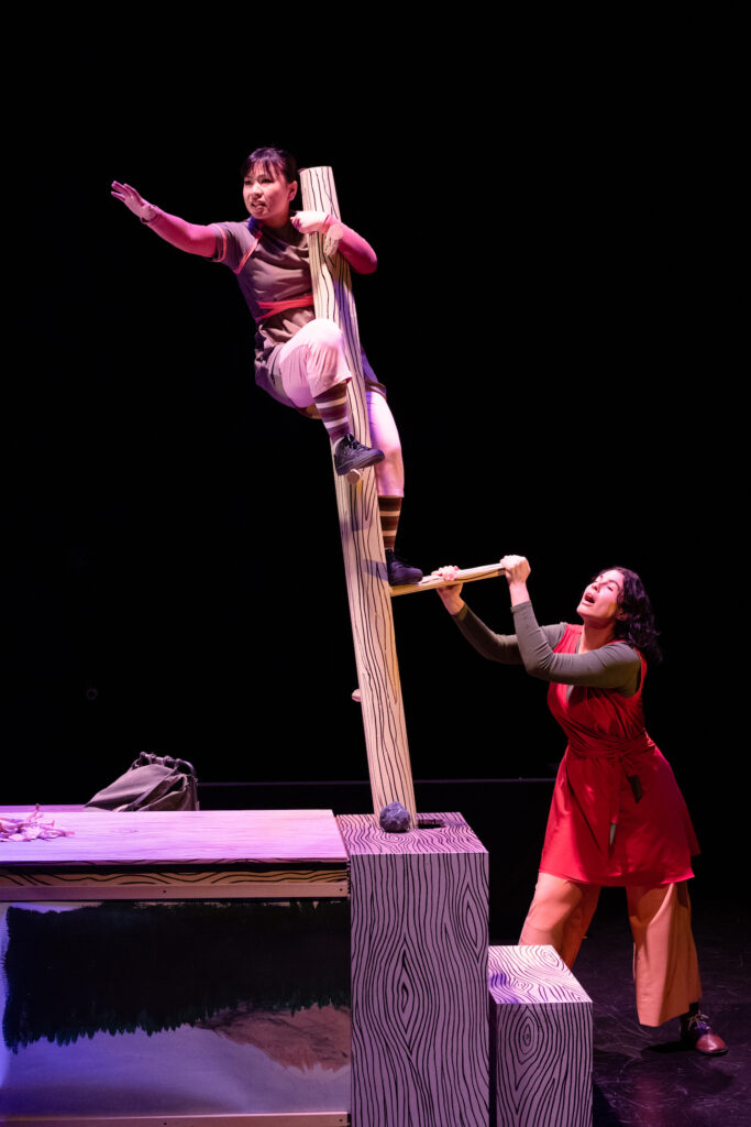 A performer clings to a wooden post, another grasps a handle to manipulate it.