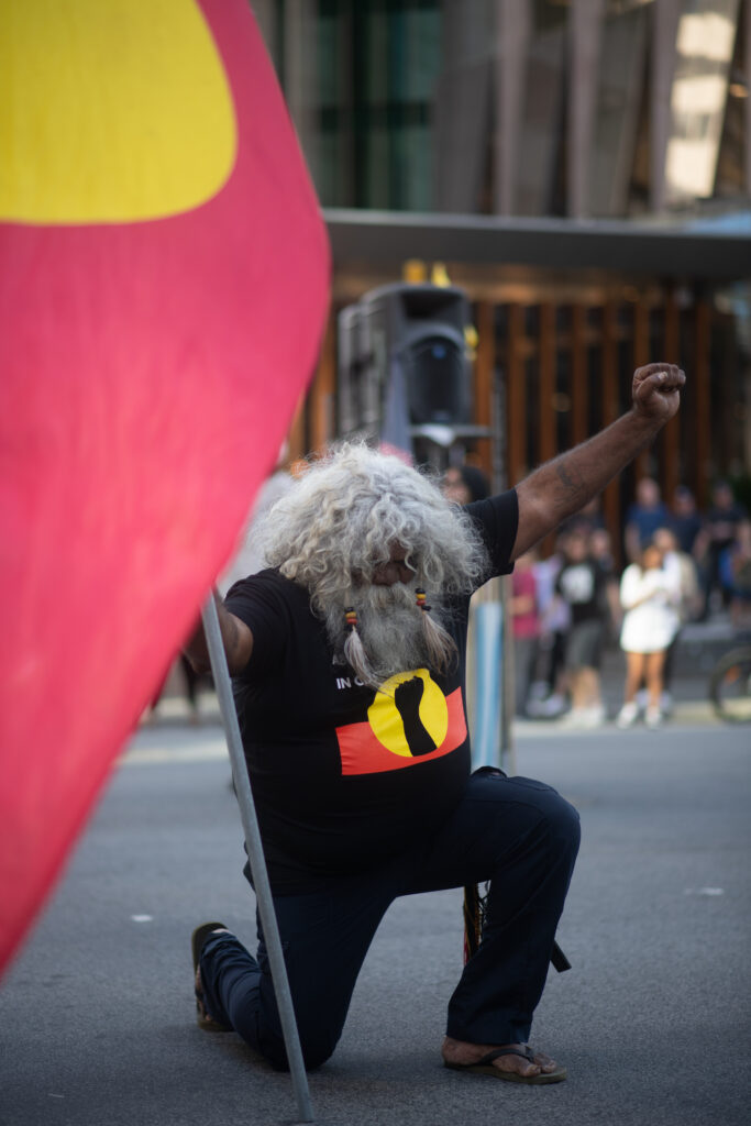 A First Nations man wearing a black t-shirt with the Aboriginal flag and a raised black fist in the centre, kneels and raises one fist.
