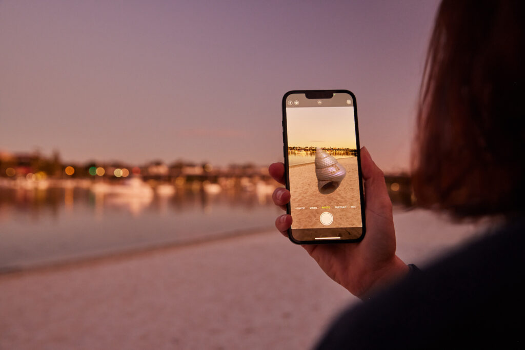 A person holds up a mobile phone as though taking a photo of a beach but there is a shell on the screen that isn't in the actual scene.